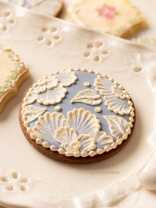 A round cookie decorated with blue icing and white brush embroidered flowers. The cookie is on a platter with an eyelet lace design around the edge.