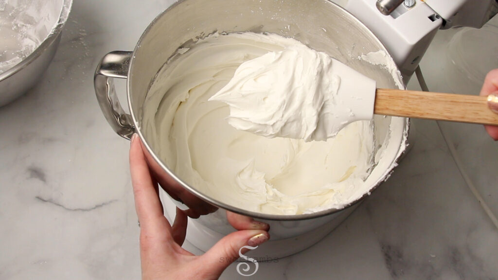 A mixing bowl full of white royal icing. A spatula is above the bowl covered in a dollop of icing. A hand is holding the bowl on the left side.