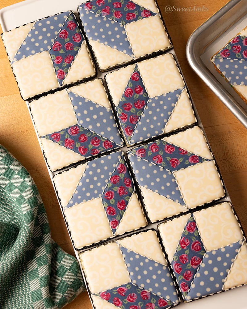 A set of square shaped cookies decorated to look like a patchwork quilt in muted colors. The cookies are arranged vertically on a platter surrounded by a dish towel and the corner of a sheet tray.