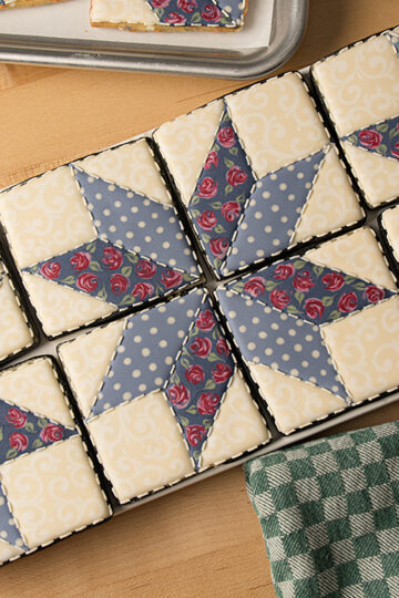 A set of square shaped cookies decorated to look like a patchwork quilt in muted colors. The cookies are arranged horizontally on a platter surrounded by a dish towel and the corner of a sheet tray.
