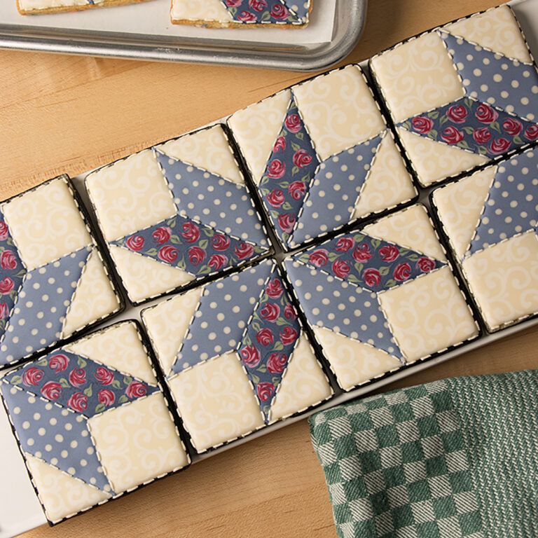 A set of square shaped cookies decorated to look like a patchwork quilt in muted colors. The cookies are arranged horizontally on a platter surrounded by a dish towel and the corner of a sheet tray.