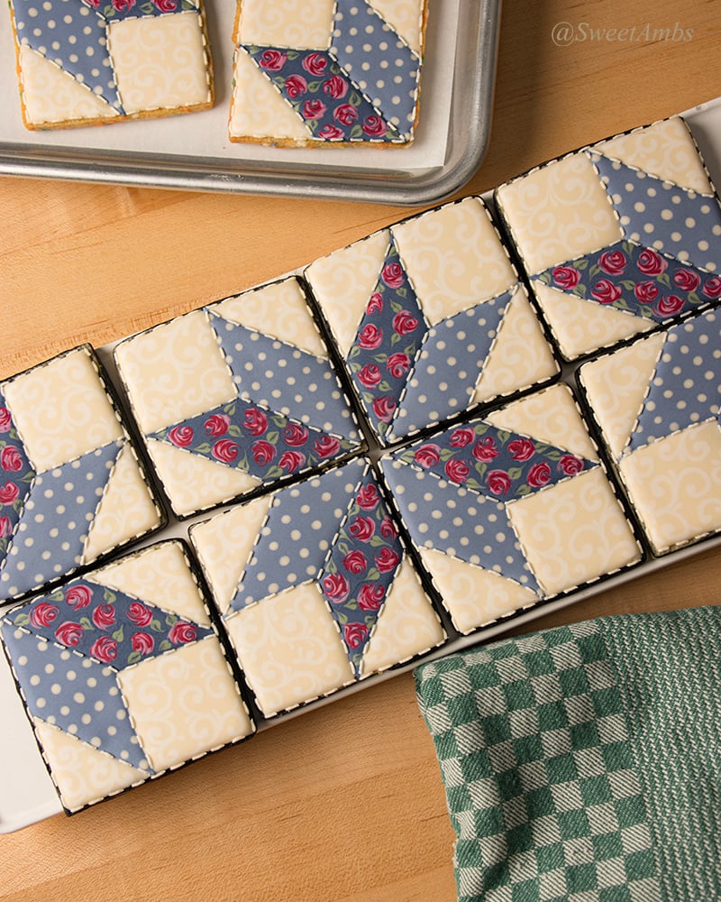 A set of square shaped cookies decorated to look like a patchwork quilt in muted colors. The cookies are arranged horizontally on a platter surrounded by a dish towel and the corner of a sheet tray.