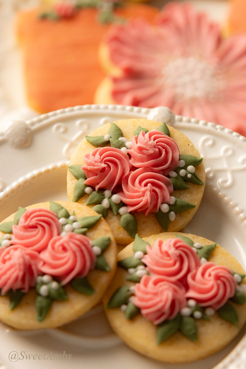 A set of 3 small round cookies decorated with swirl flowers and green leaves with small white accents on a decorative platter. There are more decorated cookies out of focus in the background.
