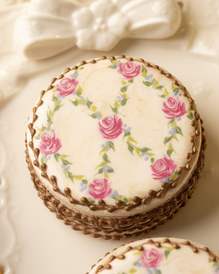 Cookies decorated to resemble vintage floral enameled makeup compacts. There are 3 cookie boxes on a plate and one of the lids is askew, revealing bronzed chocolate chips inside. The platter has an eyelet lace edge.
