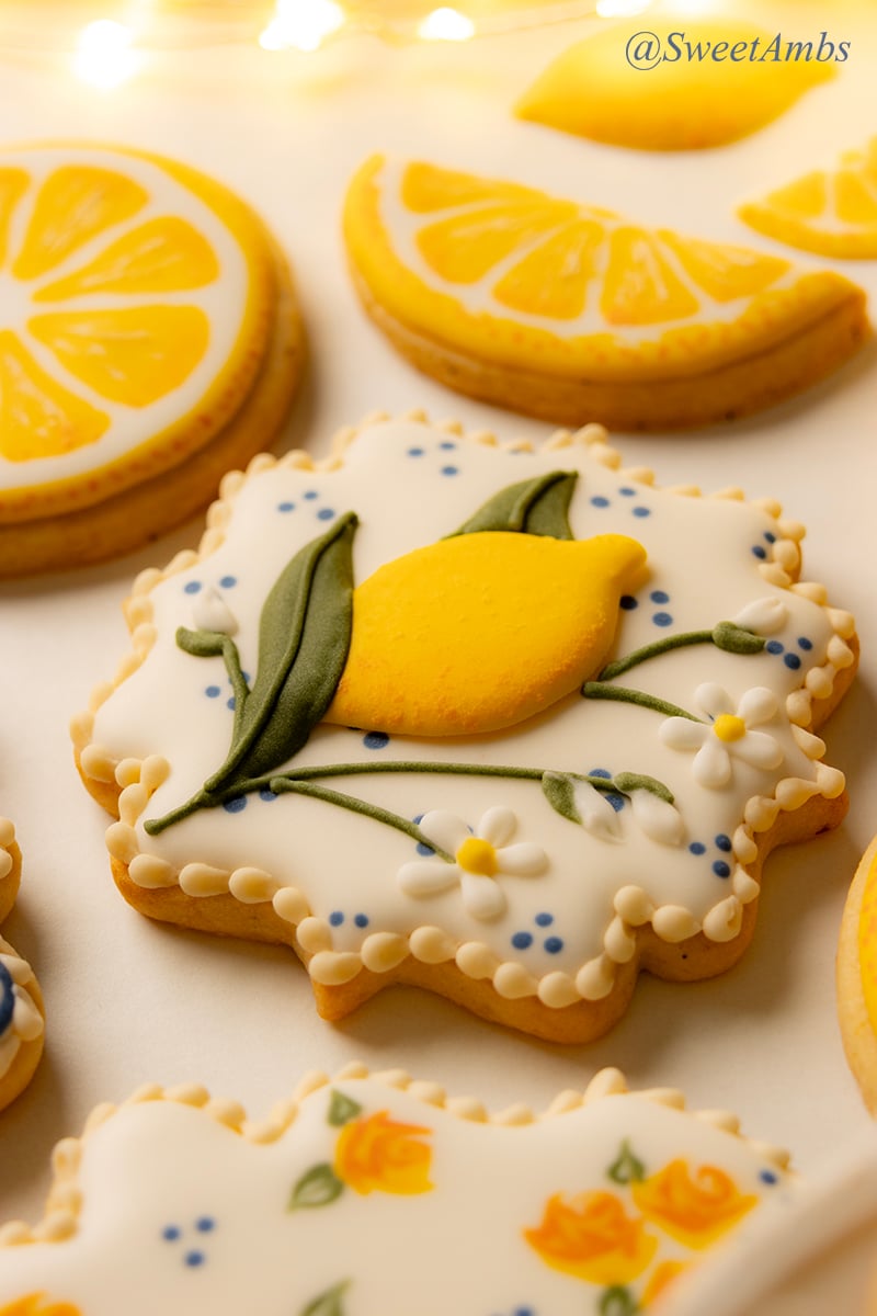 A square plaque shaped cookie decorated with a lemon branch design. There are lemon shaped cookies and fairy lights in the background.