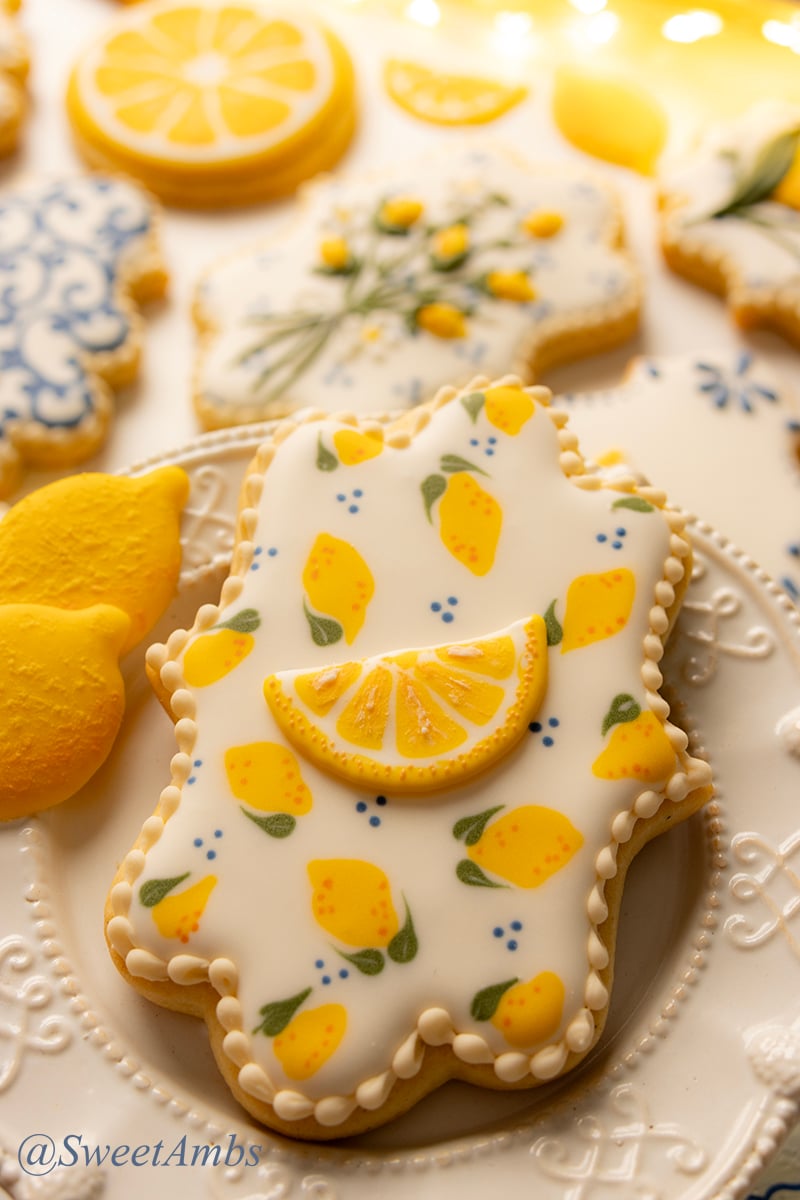 A plaque shaped cookie decorated with a lemon pattern on a decorative plate. There are more lemon themed decorated cookies out of focus in the background.