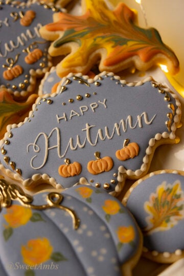 A plaque shaped cookie decorated with blue icing, small orange pumpkins, and gold details with "happy autumn" piped in the center. The cookie is surrounded by other fall decorated cookies.