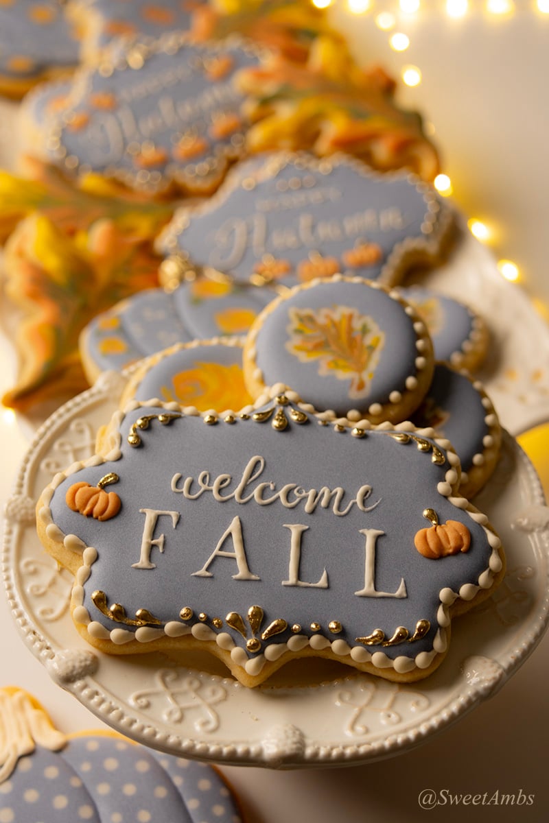 A plaque shaped cookie decorated with blue icing, small orange pumpkins, and gold details with "welcome fall" piped in the center. There are more fall cookies and fairy lights out of focus in the background.