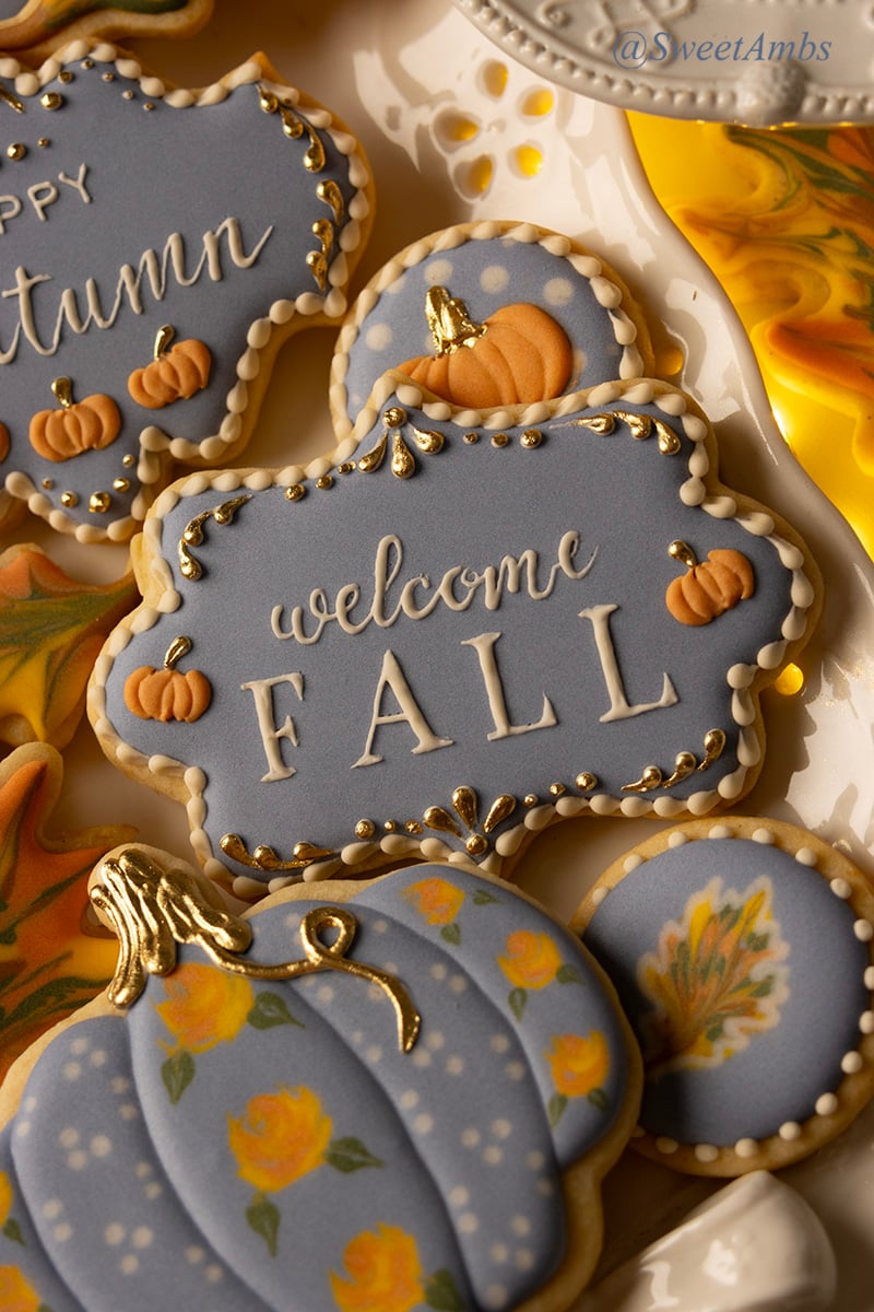 A plaque shaped cookie decorated with blue icing, small orange pumpkins, and gold details with "welcome fall" piped in the center. The cookie is surrounded by other fall decorated cookies.