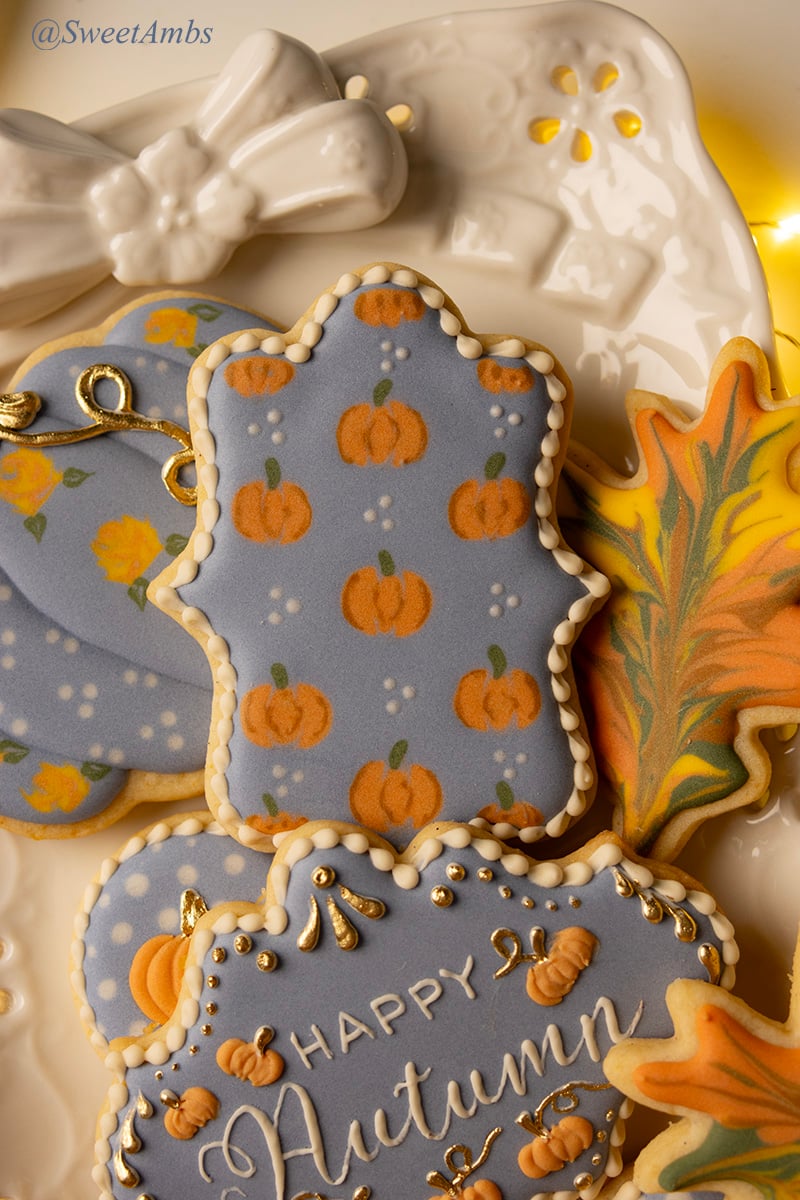 A plaque shaped cookie decorated with a wet-on-wet pumpkin design on blue icing. The cookie is on a platter with a porcelain bow and surrounded by other fall cookies.