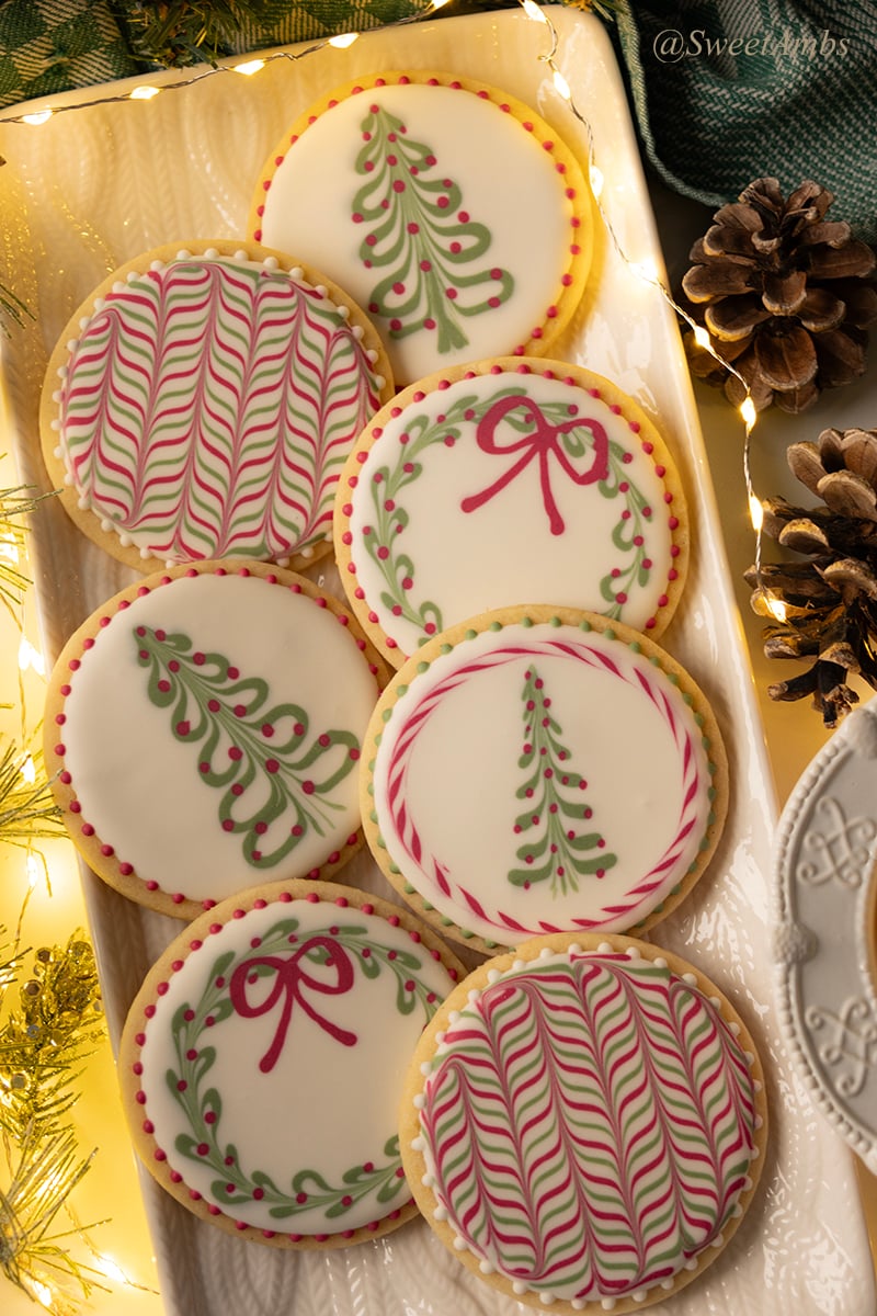 A platter of round cookies decorated with Christmas designs including a tree, a wreath, and a colorful herringbone pattern. The cookies are surrounded by fairy lights, pine cones, and a green gingham dish towel.