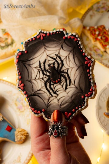 A plaque shaped cookie decorated with a spider on a spider web over a glowing white background that fades into a black border. The cookie is decorated with red and white accents and it's being held by a hand with shiny red nails with a spider ring. There are plates with fall cookies and fairy lights that are out of focus in the background.