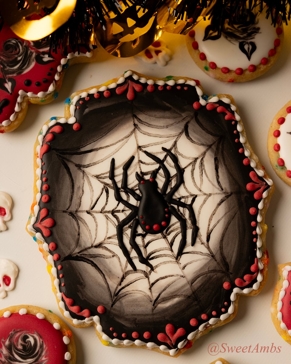 A plaque shaped cookie decorated with a spider on a spider web over a glowing white background that fades into a black border. The cookie is decorated with red and white accents and it's surrounded by other Halloween cookies, decorations, and fairy lights.