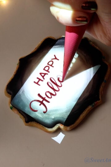 A cookie with a bright rectangular image being projected onto it and a piping bag tracing over lettering that reads Happy Halloween