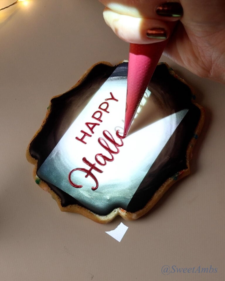A cookie with a bright rectangular image being projected onto it and a piping bag tracing over lettering that reads Happy Halloween