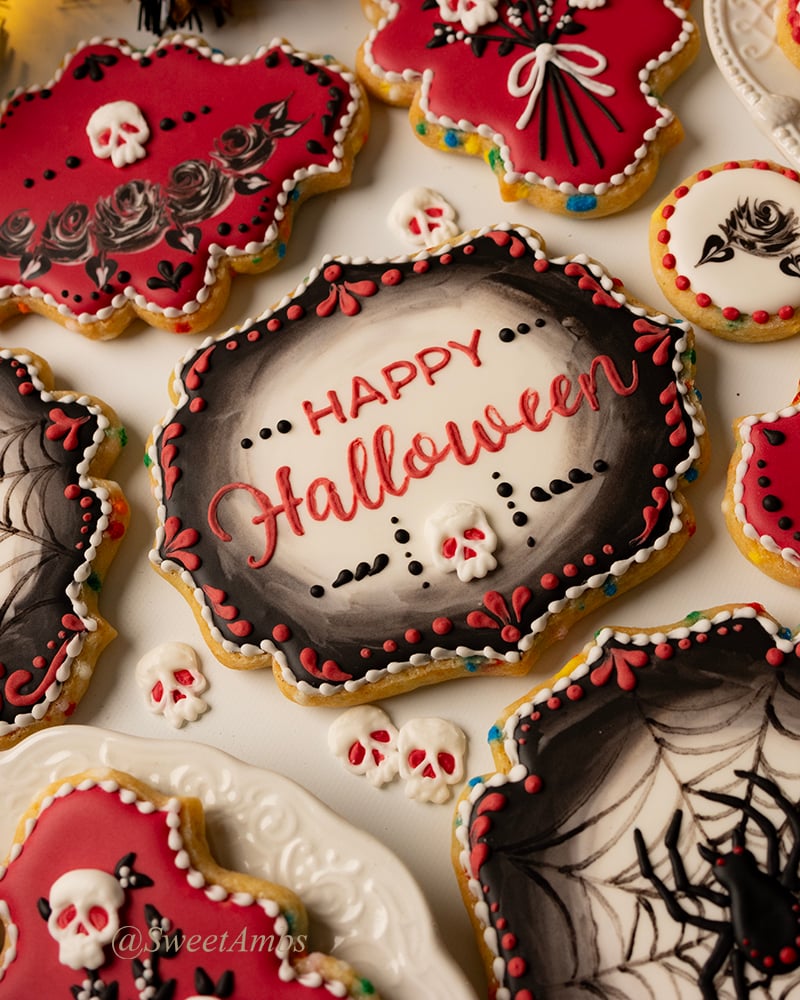 A set of plaque shaped cookies decorated with black, red, and white icing in Halloween Designs. The cookies feature skulls, a spider, roses, and Happy Halloween written across the largest plaque in the center.